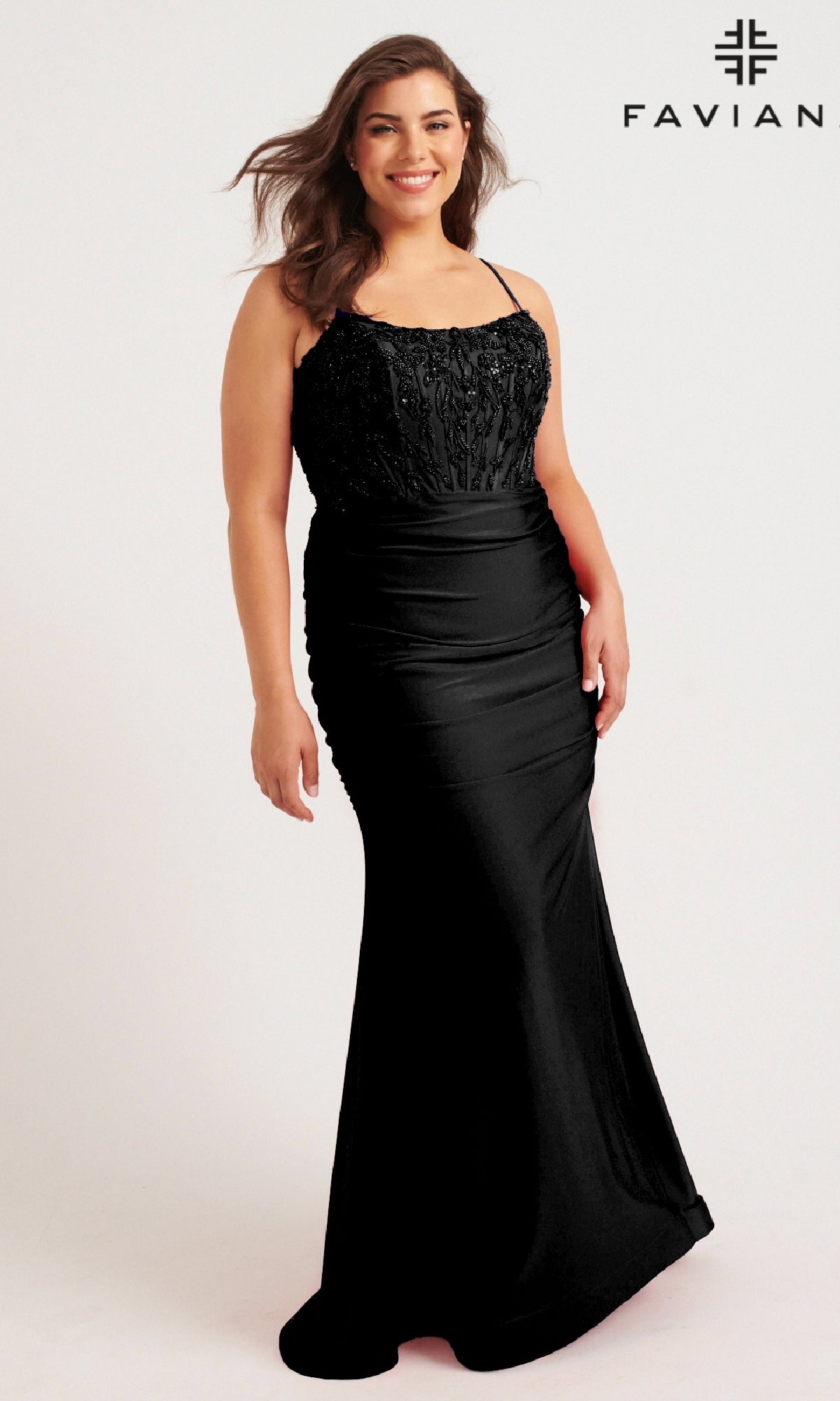 A woman in the Faviana 9569 Long Plus-Size Prom Dress—a fitted, floor-length black gown with thin straps and a beaded corset bodice—smiles against a plain background, with the “FAVIANA” logo in the top right corner.